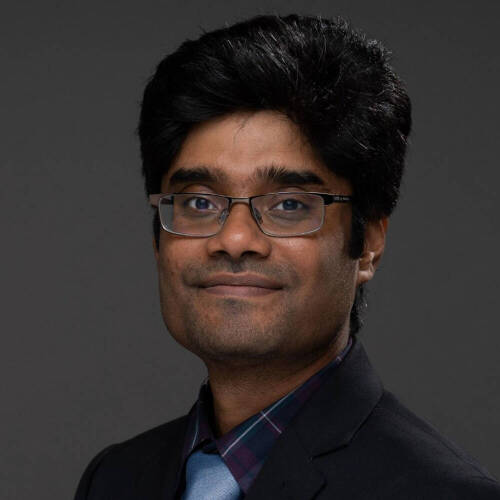 Dr. Rahat Rafiq with short, dark hair is wearing a dark suit jacket, a blue tie, a plaid shirt and a pair of glasses. The background is a plain, neutral gray. He poses for a formal portrait.